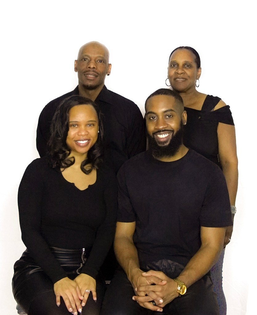 A group portrait of four people, smiling and posing together in front of a white background. They are dressed in black clothing, showcasing a friendly and welcoming atmosphere.