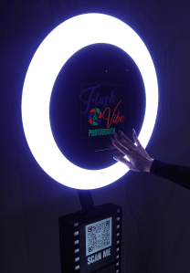 A glowing photo booth with a circular light ring and a sign that says 'Flash & Vibe Photobooth.' A hand is reaching towards the booth's touchscreen display.